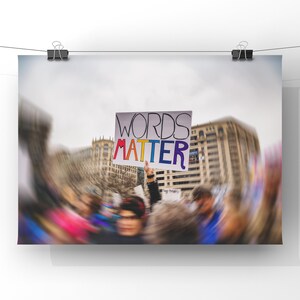 May include: A poster with the words "WORDS MATTER" in colorful letters is held up in a crowd. The sign is white with the words in a rainbow of colors. The background is blurred, suggesting a protest or rally.
