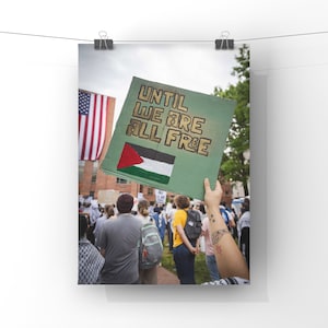 May include: A green sign with the text "UNTIL WE ARE ALL FREE" in gold lettering. The sign also features a Palestinian flag. The sign is being held up by a person with a tattoo on their arm. The sign is in front of a crowd of people.