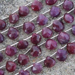 Pode incluir: Primeiro plano de fios de contas de rubi facetadas em forma de coração. As pedras preciosas vermelho-púrpura escuras são enfiadas em cordões brancos. As contas são dispostas diagonalmente sobre uma superfície cinza texturizada.