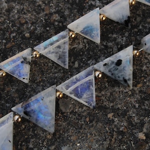 May include: Close-up of a necklace featuring triangular moonstone beads. Each translucent bead has a unique pattern of blue and black inclusions, connected by small gold-colored beads. The necklace is displayed on a textured, dark gray surface.