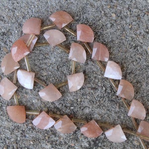 Può includere: Una collana di perline di pietre preziose sfaccettate color pesca. Ogni perla ha la forma di una mezzaluna, infilata con metallo color oro. Le perle sono disposte su una superficie grigia e strutturata.