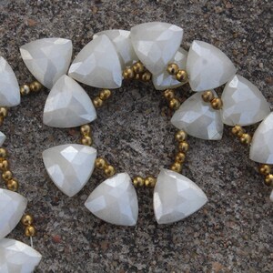 Op de afbeelding: Een ketting met facetgeslepen, driehoekige, gebroken witte kralen. De kralen zijn geregen met kleine, goudkleurige kralen. De ketting wordt getoond op een getextureerd, grijs oppervlak. Het ontwerp is elegant en geometrisch.