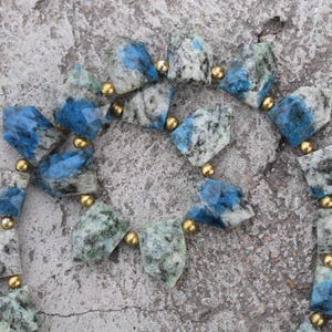 Könnte beinhalten: Eine Halskette mit facettierten, unregelmäßig geformten Steinen in Blau-, Grau- und Grüntönen, die durch kleine goldfarbene Perlen verbunden sind. Die Halskette liegt auf einer strukturierten, grauen Oberfläche.
