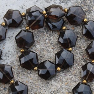 Könnte beinhalten: Eine Halskette mit facettierten, dunkelbraunen sechseckigen Perlen. Die Perlen sind mit kleinen goldfarbenen Perlen aufgefädelt. Die Halskette wird auf einer strukturierten, hellgrauen Oberfläche präsentiert.