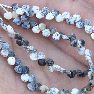 Peut inclure: Brins de perles d'opale dendritique en forme de goutte. Les perles sont blanches avec des mouchetures grises et noires, enfilées avec du fil argenté. Les perles sont présentées sur un fond de couleur chair.