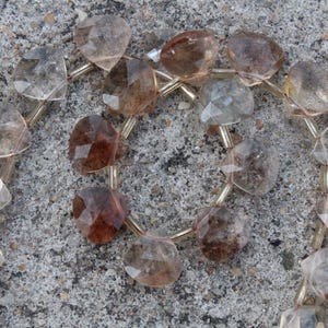 May include: A necklace featuring heart-shaped, faceted gemstones in shades of brown and clear. The beads are strung together with gold-colored wire, creating a delicate and elegant design. The necklace is displayed on a textured, gray surface.