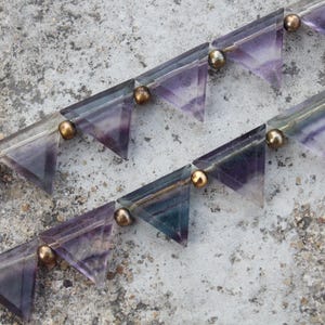 May include: Close-up of two strands of gemstone beads. Each strand features triangular, translucent fluorite beads in shades of purple and green, connected by small, gold-colored metal beads. The background is a textured, gray surface.