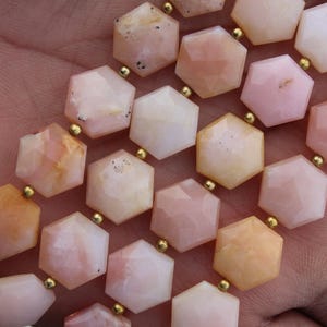 Puede incluir: Primer plano de un collar con cuentas hexagonales en tonos melocotón, rosa y crema. Las cuentas están ensartadas con conectores dorados. Las piedras preciosas tienen una superficie facetada, creando un efecto brillante.