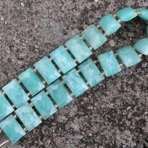 Peut inclure: Deux rangées de perles rectangulaires facettées en amazonite, dans des tons turquoise et vert d'eau. Les perles sont reliées par des entretoises dorées, pour un collier ou un bracelet. Le fond est une surface rugueuse et texturée.