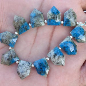 Peut inclure: Un chapelet de perles de k2 azurite facettées. Les perles sont un mélange de bleu, de gris et de blanc, de forme géométrique. Chaque perle est séparée par une petite perle argentée. Les perles sont enfilées sur une ficelle bleu clair.
