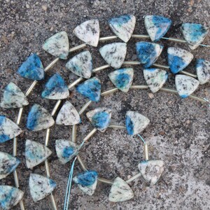 Peut inclure: Des rangées de perles triangulaires en jaspe K2. Les perles sont un mélange de blanc, de noir et de bleu, avec un aspect moucheté. Les perles sont enfilées sur un fil fin de couleur or.
