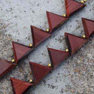 Op de afbeelding: Een ketting met driehoekige, roodbruine houten kralen verbonden door kleine, goudkleurige bollen. Het geometrische ontwerp is gerangschikt in een herhalend patroon, waardoor een uniek accessoire ontstaat.