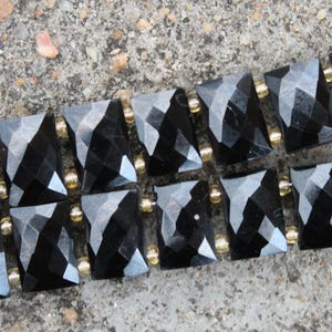 Può includere: Primo piano di un braccialetto nero sfaccettato con dettagli color oro. Il braccialetto presenta pietre nere lucide di forma quadrata, collegate da piccole perle dorate e maglie in metallo dorato. Il gioiello è appoggiato su una superficie grigia strutturata.