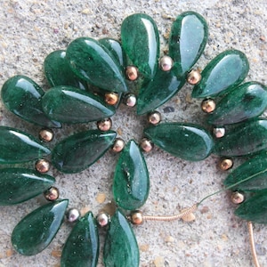 Puede incluir: Una colección de cuentas de piedras preciosas de color verde oscuro en forma de lágrima, dispuestas en un patrón circular e intercaladas con pequeñas cuentas espaciadoras redondas de tono dorado metálico, ensartadas en un hilo fino y flexible.