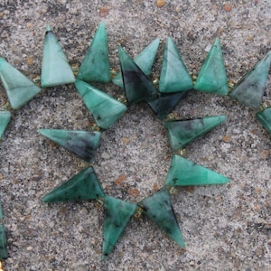Op de afbeelding: Een ketting met driehoekige, smaragdgroene edelstenen. De stenen zijn in een cirkelvormig patroon gerangschikt, verbonden door goudkleurige kralen. De ketting wordt getoond op een getextureerd, grijs oppervlak.