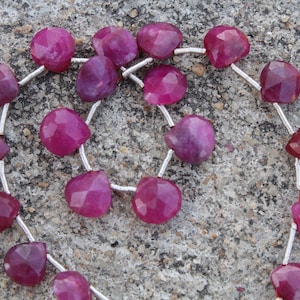 May include: A strand of faceted, heart-shaped ruby beads. The deep magenta gemstones are strung on a silver-colored wire. The beads are displayed on a textured, gray surface.