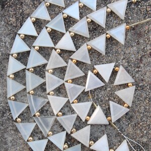 Peut inclure: Gros plan sur une guirlande décorative composée de perles triangulaires blanches translucides. Chaque triangle est relié par une petite perle dorée. La guirlande est disposée en cercle.