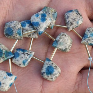 Può includere: Una fila di perline di pietra K2. Ogni perla ha una forma unica, con una miscela di tonalità bianche, grigie e blu. Le perle sono collegate da aste metalliche color oro e infilate su un filo azzurro, ideali per la creazione di gioielli.