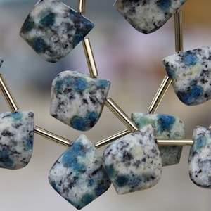 Può includere: Primo piano di una collana con perline di pietra K2. Le perline sono un mix di bianco, grigio e nero, con accenti blu. Le perline sono infilate con aste di metallo color oro, creando un gioiello unico.