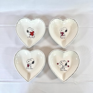 May include: Four heart-shaped white ceramic dishes with black trim. Each dish features a Snoopy character holding a red heart. The dishes are arranged on a white surface.
