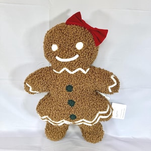 Puede incluir: Un muñeco de jengibre de peluche marrón con un lazo rojo, detalles blancos y botones verdes. El muñeco de jengibre tiene una cara sonriente con ojos blancos y una boca blanca. El peluche está diseñado para parecer una galleta de jengibre.