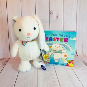 May include: A white plush bunny with a bow tie holds a colorful Easter basket. The bunny is standing next to a book titled "Hopper Saves Easter" with a cartoon bunny on the cover.