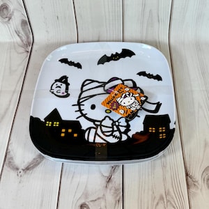 May include: A white plastic square plate with a black and white Halloween design featuring Hello Kitty dressed as a mummy. The plate has bats and houses with windows. The plate is stacked on top of another plate. The plate has a Hello Kitty Halloween sticker attached to it.