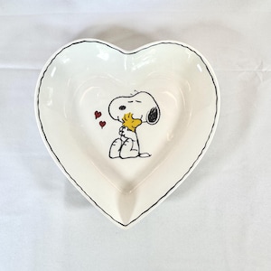 May include: A heart-shaped, white ceramic dish with a black outline. The dish features a cartoon illustration of Snoopy hugging Woodstock, with three small red hearts. The dish is likely used for serving or display.