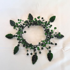 May include: A handcrafted, dark green beaded crown with dark green leaves and berries. The crown is made of dark green beads and leaves, and is arranged in a circular shape. The leaves are a darker shade of green.