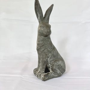 Könnte beinhalten: Eine graue Hasenstatue mit langen Ohren und einer sitzenden Pose. Die Skulptur hat eine strukturierte Oberfläche und befindet sich vor einem weißen Hintergrund. Die Hasenfigur ist ein dekorativer Artikel.