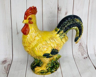 Peut inclure: Une figurine de coq en céramique avec des détails peints en jaune, vert et noir. Le coq a une crête et une barbe rouges et une queue noire.