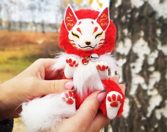May include: A red and white plush fox with a golden mask and a bell around its neck. The fox is sitting in a person's hands.