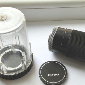 Può includere: Un obiettivo per fotocamera vintage con custodia protettiva e tappo. L'obiettivo è nero con dettagli argentati e segni di misurazione. La custodia trasparente ha una parte superiore argentata e una base nera. Il tappo dell'obiettivo è nero con testo bianco.