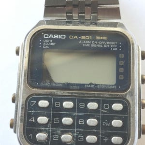 May include: A vintage Casio CA-901 digital watch with a silver-tone metal band and a rectangular face. The watch features a calculator keypad and a digital display. The watch band is approximately 0.75 inches wide.