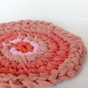 May include: A round rug in shades of pink and coral. The rug is made from braided fabric strips, creating a textured surface. The design features concentric circles of varying colours, with a darker pink border.