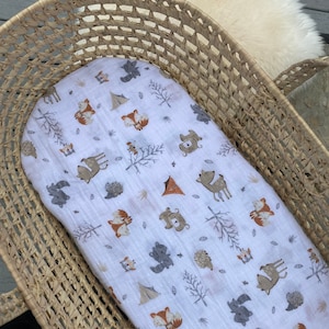 May include: A white muslin baby blanket with a woodland animal print featuring foxes, deer, squirrels, bears, and hedgehogs. The blanket is in a woven wicker bassinet.