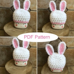 May include: A crochet bunny hat with pink ears and a white body. The hat is sitting on a white ceramic mug with a happy face.
