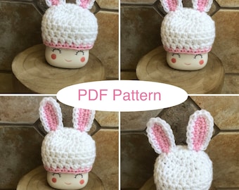 May include: A crochet bunny hat with pink ears and a white body. The hat is sitting on a white ceramic mug with a happy face.