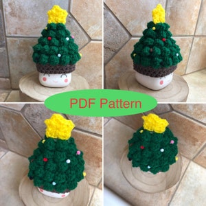 May include: A crochet Christmas tree hat in green with a yellow star on top. The hat is on a white mug with a brown rim and a smiling face. The mug is sitting on a wooden coaster.