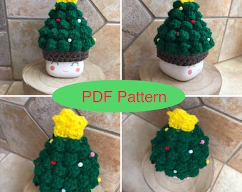 May include: A crochet Christmas tree hat in green with a yellow star on top. The hat is on a white mug with a brown rim and a smiling face. The mug is sitting on a wooden coaster.