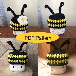 May include: Crochet bee hat pattern for a mug cozy. The pattern is available as a PDF download. The hat is black with yellow stripes and has a white flower on top. The hat is on a white mug with a smiley face.
