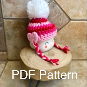 May include: A pink and red crocheted hat with a white pom pom and a pink heart on the side. The hat is on a white mug with a happy face. The mug is sitting on a wooden stand. PDF Pattern.