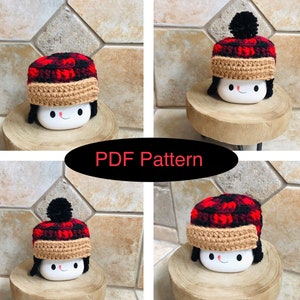 May include: A crochet pattern for a red and black plaid hat with a brown brim and a black pom-pom on top. The hat is sitting on a white ceramic mug with a happy face.