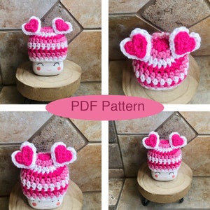 May include: A pink and white crocheted hat with heart-shaped earflaps. The hat is sitting on a small wooden pedestal. The text "PDF Pattern" is visible in the image.