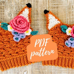 May include: Two crocheted fox hats with flower embellishments. The hats are orange with white fox ears and feature a variety of crocheted flowers in pink, purple, white, and green. The text "PDF pattern" is written on a peach-colored background.