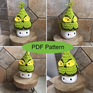 May include: A crochet Grinch-inspired mug cosy in green with yellow eyes and a black mouth. The cosy is on a white mug with a smiling face. The mug is on a wooden coaster.