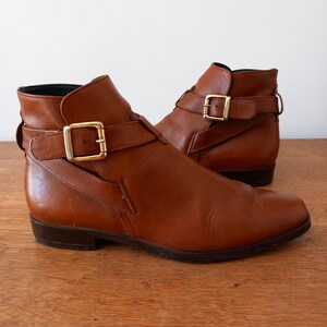 May include: A pair of brown leather ankle boots with gold-coloured buckle details. The boots have a low heel and a classic design, suitable for various outfits. The leather appears smooth and well-maintained.