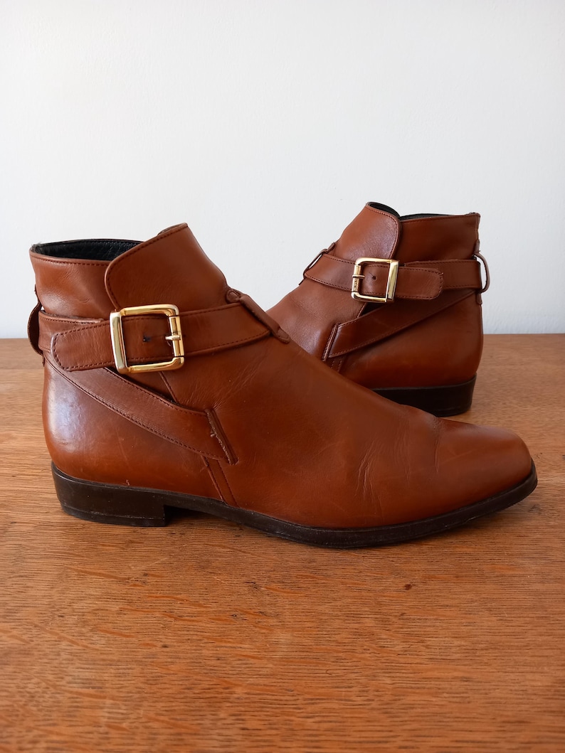 May include: A pair of brown leather ankle boots with gold-coloured buckle details. The boots have a low heel and a classic design, suitable for various outfits. The leather appears smooth and well-maintained.