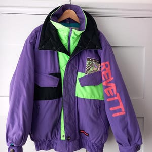 May include: A purple, black, and neon green jacket featuring a high collar, a front zipper, and two chest pockets. The word REVETTI is printed in large pink letters on the left sleeve, with a small graphic patch above the left chest pocket.