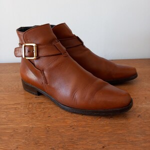 May include: A pair of brown leather ankle boots with a buckle closure and a gold-coloured buckle. The boots have a low heel and a rounded toe. The boots are displayed on a wooden surface.